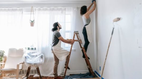 A couple repainting the wall, women painting on the ladder, man holding the ladder steady.