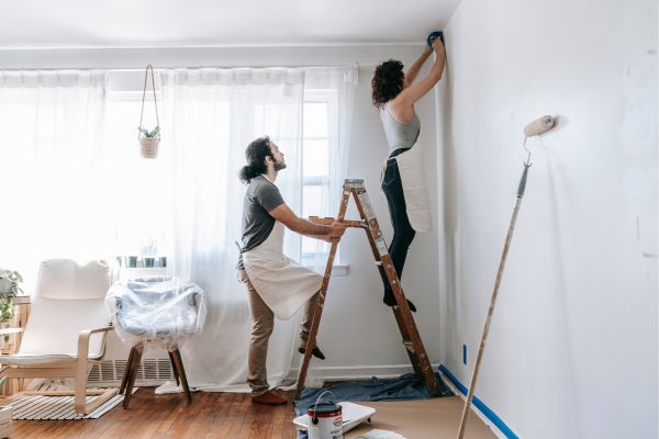 A couple repainting the wall, women painting on the ladder, man holding the ladder steady.